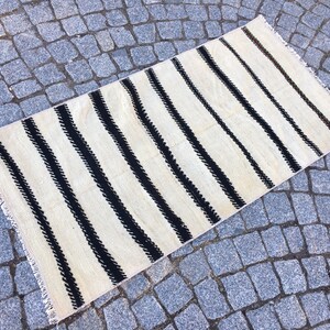 K&ouml;nnte beinhalten: Ein wei&szlig;-schwarz gestreifter Teppich mit Fransenrand. Der Teppich hat ein schlichtes Design mit schwarzen Streifen auf wei&szlig;em Hintergrund.