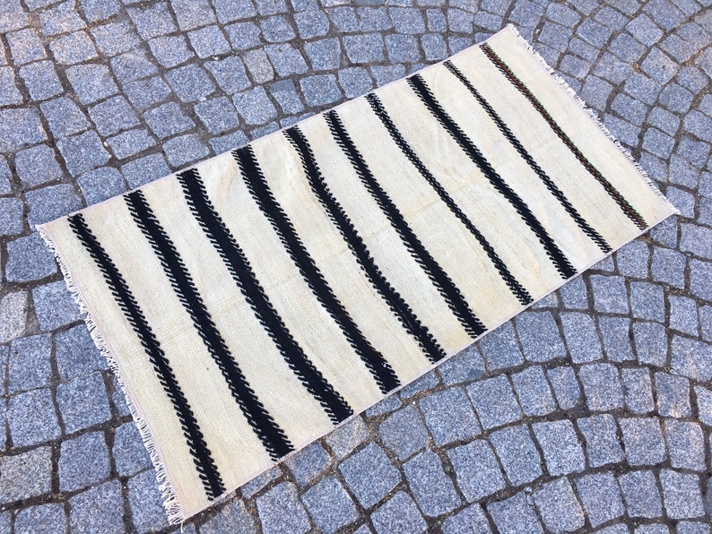 K&ouml;nnte beinhalten: Ein wei&szlig;-schwarz gestreifter Teppich mit Fransenrand. Der Teppich hat ein schlichtes Design mit schwarzen Streifen auf wei&szlig;em Hintergrund.