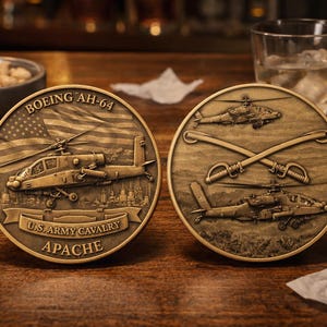 May include: Two bronze-coloured Apache helicopter coins. One coin features a helicopter, American flag, and text "BOEING AH-64" and "U.S. ARMY CAVALRY APACHE". The other coin shows crossed swords and a helicopter. The coins are on a wooden surface.