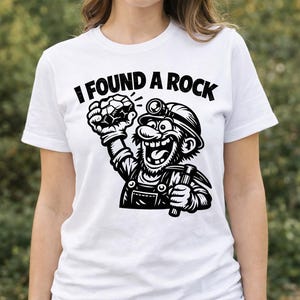 Puede incluir: Camiseta blanca con un dibujo en negro de un minero de dibujos animados sosteniendo una gema y un pico, con el texto "I FOUND A ROCK". La camiseta la lleva una persona.
