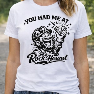 Puede incluir: Camiseta blanca con un gráfico de un minero de dibujos animados sosteniendo un diamante, el texto dice "YOU HAD ME AT" arriba y "Rock Hound" abajo. El minero lleva un casco y sonríe.