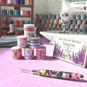 May include: A selection of sewing accessories, including thread spools, a seam ripper, and a Singer sewing machine. The spools feature diverse patterns and colours, and the seam ripper has a floral design. A sign reads "FOXGLOVE & FIELD."