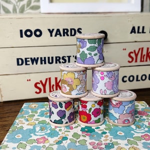 May include: Six wooden spools of thread with floral patterns. The spools are stacked in a pyramid shape. The spools are all different colours and patterns. The spools are sitting on a floral patterned fabric.