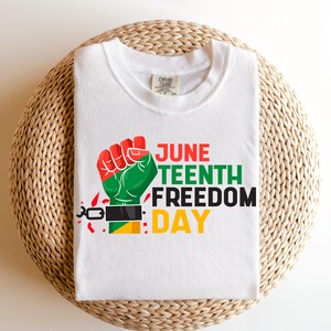 Può includere: Maglietta bianca con una grafica di un pugno alzato che si libera dalle catene. Il testo "Juneteenth Freedom Day" è stampato in rosso, verde e giallo.