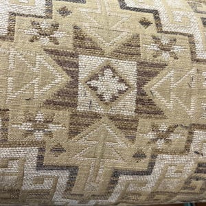 May include: Close-up of a woven textile with a geometric pattern. The design features repeating shapes in shades of beige, brown, and cream. The texture appears to be coarse, with visible threads creating a detailed surface.