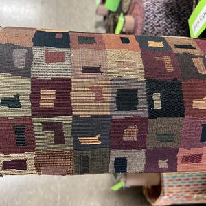 May include: Close-up of a fabric roll with a geometric pattern. The design features squares in shades of brown, burgundy, black, and tan. The fabric has a textured appearance, suggesting a woven material. The overall aesthetic is reminiscent of a vintage or rustic style.