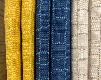May include: Close-up of four fabric swatches. The swatches are arranged vertically. The first swatch is a bright yellow with a subtle woven pattern. The second swatch is a deep yellow with a more pronounced woven pattern. The third swatch is a dark blue with a woven pattern of small squares. The fourth swatch is a light beige with a woven pattern of small squares.