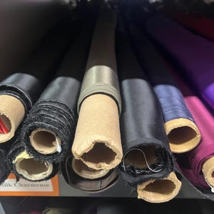 K&ouml;nnte beinhalten: Nahaufnahme von mehreren Stoffrollen in verschiedenen Farben, darunter Schwarz, Grau, Lila und Blau. Der Stoff ist aufgerollt und mit Pappr&ouml;hren befestigt. Das Etikett "Silk Charmeuse" ist unten links sichtbar.