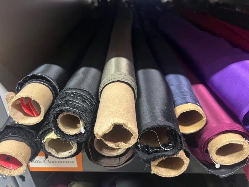 K&ouml;nnte beinhalten: Nahaufnahme von mehreren Stoffrollen in verschiedenen Farben, darunter Schwarz, Grau, Lila und Blau. Der Stoff ist aufgerollt und mit Pappr&ouml;hren befestigt. Das Etikett "Silk Charmeuse" ist unten links sichtbar.