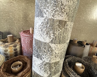 May include: A large roll of textured carpet with a geometric pattern in shades of gray and white. The carpet is rolled up, revealing its thickness and design. Other rolls of patterned carpets are visible in the background.
