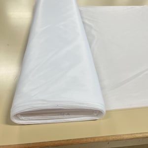 May include: A white fabric roll, folded and sitting on a wooden table. The fabric is smooth and appears to be a lightweight material.