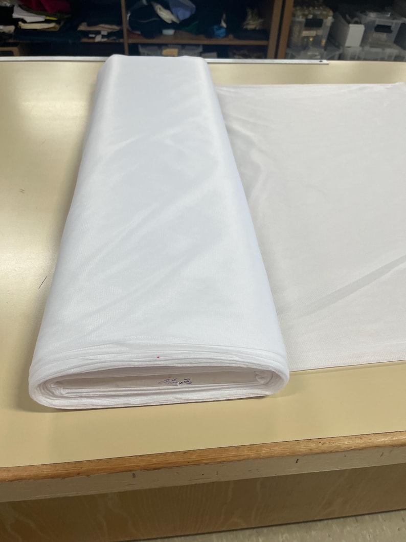 May include: A white fabric roll, folded and sitting on a wooden table. The fabric is smooth and appears to be a lightweight material.
