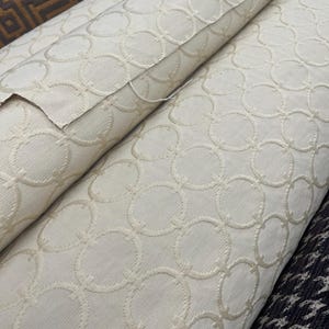 May include: Rolled fabric with a cream-colored background and a repeating pattern of interlocking circles. The fabric has a textured appearance, with the circles slightly raised. Other fabrics with geometric patterns are visible in the background.