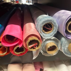 K&ouml;nnte beinhalten: Rollen von Stoff in verschiedenen Farben, darunter Blau, Rosa und Lila, sind in einem Regal gestapelt. Der Stoff ist aufgerollt und mit braunem Karton befestigt.