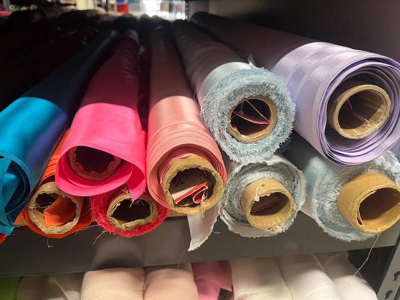 K&ouml;nnte beinhalten: Rollen von Stoff in verschiedenen Farben, darunter Blau, Rosa und Lila, sind in einem Regal gestapelt. Der Stoff ist aufgerollt und mit braunem Karton befestigt.