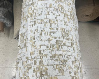 May include: A rolled-up fabric bolt featuring a white background with a gold and tan geometric pattern. The fabric has a textured appearance and a woven border. The bolt is displayed vertically.
