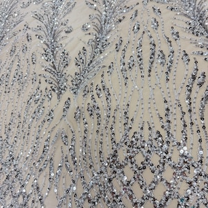 May include: Close-up of a silver sequined fabric with a delicate, flowing design. The sequins are arranged in a pattern of feathery shapes and flowing lines, creating a shimmering, textured surface. The fabric has a neutral background.