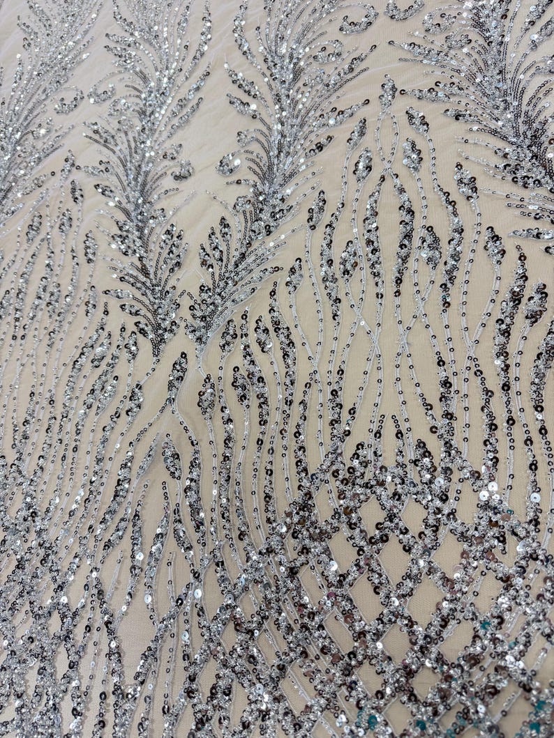 May include: Close-up of a silver sequined fabric with a delicate, flowing design. The sequins are arranged in a pattern of feathery shapes and flowing lines, creating a shimmering, textured surface. The fabric has a neutral background.