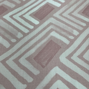 May include: Close-up of a fabric with a repeating geometric pattern. The design features light pink and white angular shapes, creating a textured, abstract design. The fabric appears to be cotton or a similar material.