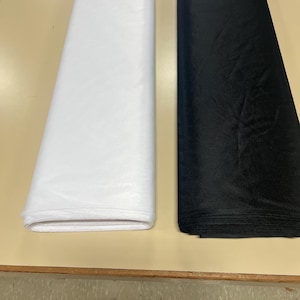 May include: Two rolls of fabric, one white and one black, are laid out on a wooden surface. The fabric is sheer and has a fine mesh texture.