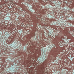 May include: Close-up of a fabric with a repeating paisley pattern. The background is a muted reddish-brown color, with a lighter, almost teal, paisley design overlaying it. The pattern is detailed and ornate.