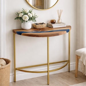 Può includere: Un tavolino a mezzaluna con piano in legno e gambe in metallo dorato. Il piano del tavolo presenta un intarsio in resina blu. Un vaso bianco con fiori, un cestino intrecciato e libri sono esposti sul tavolo. Uno specchio rotondo dorato è appeso sopra.