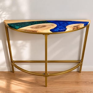 Peut inclure: Une console en demi-lune avec un plateau en bois à bord brut incrusté de résine époxy bleue et verte. La table a un cadre en métal doré avec une base incurvée et des pieds droits. La table est contre un mur blanc.
