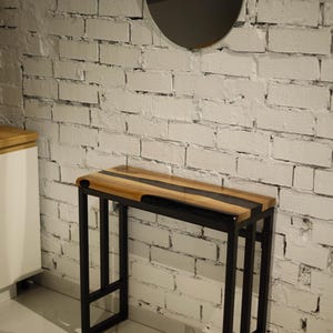 Può includere: Un tavolo consolle in legno con struttura in metallo nero. Il piano è in legno chiaro con una striscia scura al centro. Il tavolo è rettangolare e dal design moderno. Le gambe sono nere e angolari.