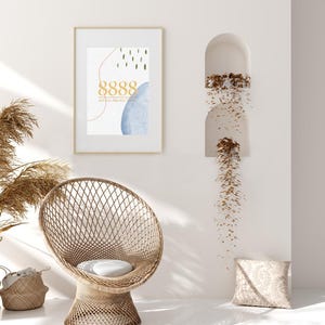 Peut inclure: Une impression encadrée avec un design abstrait bleu et blanc. L'impression présente le nombre 8888 et le texte "Divine Protection". L'impression est accrochée à un mur blanc avec un fauteuil en osier et un coussin au premier plan.