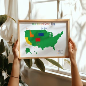 Op de afbeelding: Een ingelijste kaart van de Verenigde Staten met kleurrijke blokken die reislocaties vertegenwoordigen. De tekst "Our Travel Story Map" staat bovenaan. De kaart wordt voor een raam gehouden.
