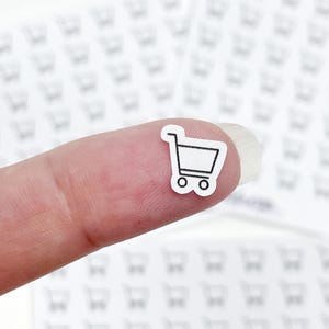 May include: A tiny white sticker with a black outline of a shopping cart is held on a finger. The background is filled with similar shopping cart stickers. The sticker is a simple, minimalist design.