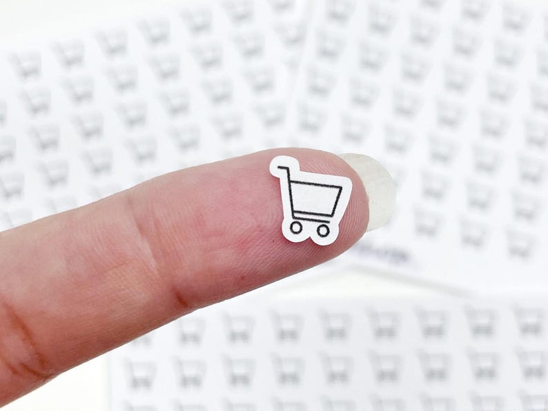 May include: A tiny white sticker with a black outline of a shopping cart is held on a finger. The background is filled with similar shopping cart stickers. The sticker is a simple, minimalist design.