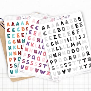 Può includere: Tre fogli di adesivi con lettere dell'alfabeto colorate. Le lettere sono in vari colori, tra cui rosso, verde acqua, arancione, viola e grigio. Gli adesivi sono su sfondi bianchi con un motivo a griglia. Il marchio "Ady Shop" è visibile.