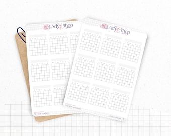 Op de afbeelding: Twee witte maandelijkse trackerstickervellen met een rasterpatroon, elk met 12 kalenderrasters. De stickers hebben de tekst "Ady Shop" en "Monthly trackers". Een bruin papier en een blauwe paperclip staan op de achtergrond.