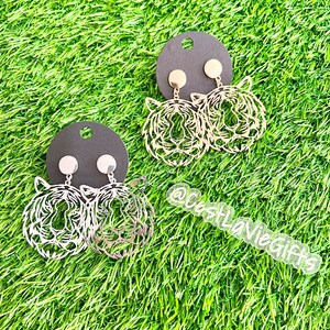 May include: Two pairs of gold tiger head earrings on black card stock. The earrings are on a green grass background. The text "@CostLaVieGifts" is visible in the lower right corner of the image.