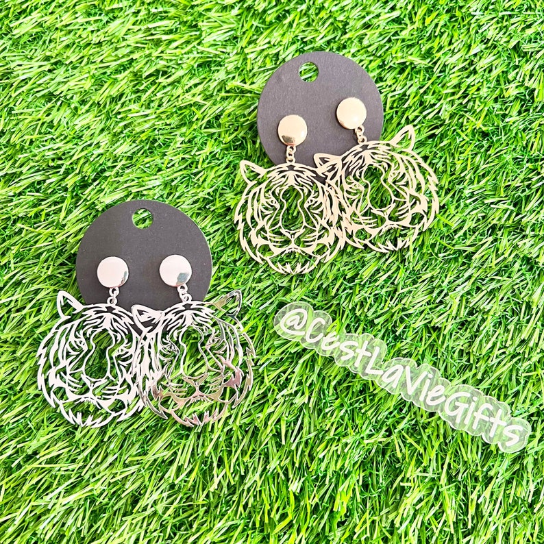 May include: Two pairs of gold tiger head earrings on black card stock. The earrings are on a green grass background. The text "@CostLaVieGifts" is visible in the lower right corner of the image.