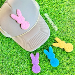 May include: A beige trucker cap with a pink bunny motif, accompanied by three more bunny patches in purple, blue, and yellow. The patches have a textured, fuzzy look. The items are set on artificial green grass.