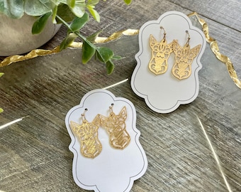 Puede incluir: Dos pares de pendientes colgantes de metal dorado, cada uno con un diseño detallado de calado en forma de cabeza de perro con orejas puntiagudas. Los pendientes se presentan sobre dos tarjetas de cartón blanco con bordes festoneados.