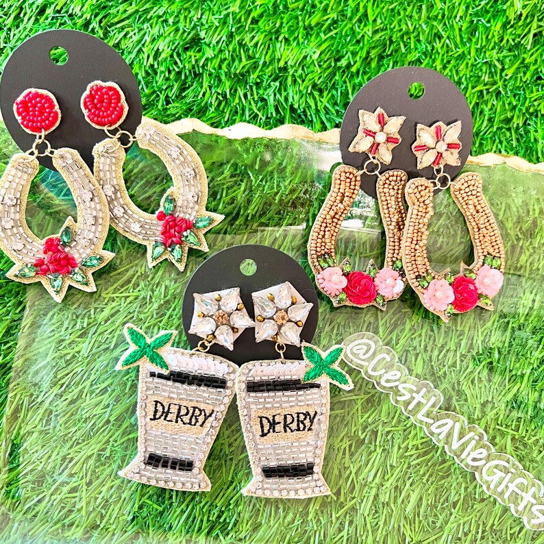 May include: Four pairs of beaded earrings with a horseshoe, flower, and cocktail glass design. The earrings are made with gold, white, and red beads. The earrings are on a white background.