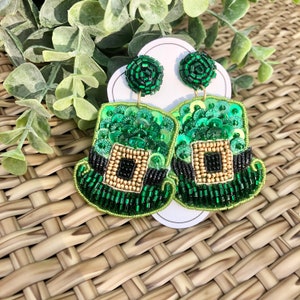 以下が含まれることがあります： 緑と金のビーズでできた、レプリコーンの帽子型のイヤリングのペア。イヤリングは緑色のスパンコールと黒いビーズで飾られています。