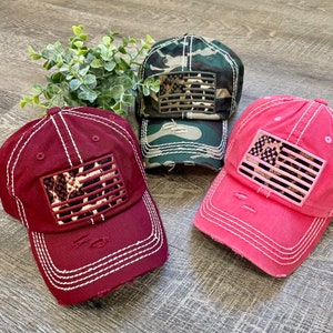 May include: Three distressed baseball caps with a leopard print American flag patch. The caps are in maroon, pink, and camouflage.