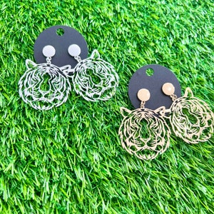May include: Two pairs of tiger face earrings. One pair is silver and the other is gold. The earrings are on black card stock.