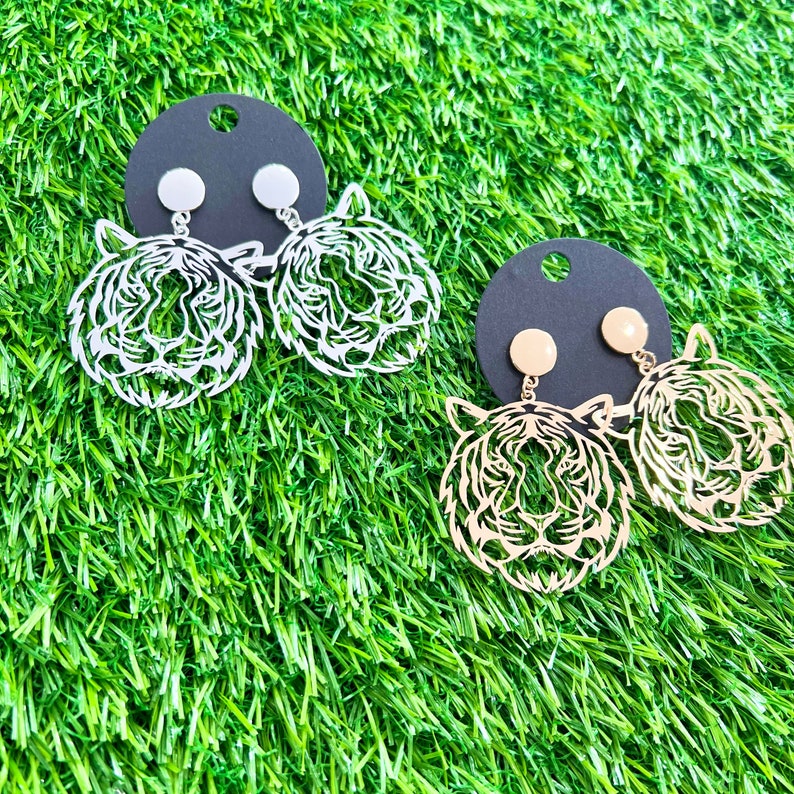 May include: Two pairs of tiger face earrings. One pair is silver and the other is gold. The earrings are on black card stock.