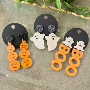 May include: Three pairs of Halloween-themed earrings. One pair features a ghost charm with three orange pumpkin charms. Another pair features a ghost charm with three orange "BOO" charms. The last pair features black glitter charms with three orange pumpkin charms.