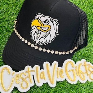 May include: Black trucker hat with a white and yellow eagle patch. A gold-colored chain with clear rhinestones is attached to the hat. The words "C'est La Vie Gifts" are displayed in gold lettering on a white background.