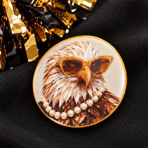 May include: A round pin featuring a detailed illustration of a bald eagle wearing amber-colored cat-eye sunglasses and a single strand of white pearl beads around its neck. The eagle has brown and white feathers and a yellow beak, set in a gold frame.