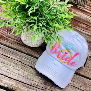 May include: A light blue distressed baseball cap with the word "bride" embroidered in pink with a gold ring and a blue diamond.