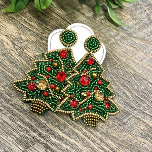 Peut inclure: Boucles d'oreilles en forme de sapin de Noël, vertes et perlées, avec des accents rouges et dorés. Les boucles d'oreilles sont ornées de perles rouges et dorées. Elles sont présentées sur une carte blanche.