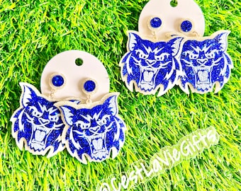 May include: A pair of blue and white glitter earrings featuring a wildcat design. The earrings are on a white background with a pink border. The text "@westlaviegifts" is visible in the bottom right corner of the image.