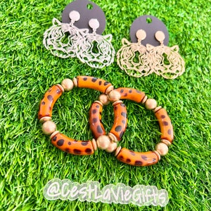 May include: Two pairs of gold tiger-shaped earrings and two brown and gold bracelets with a leopard print design. The jewelry is displayed on a green grassy surface.